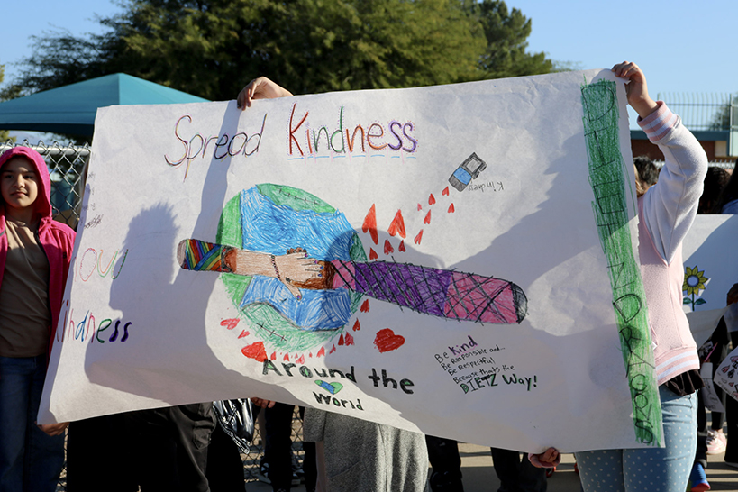 Students hold up a hand-drawn banner with the words Spread Kindness Around the World and a drawing of the Earth with two hands reaching across it