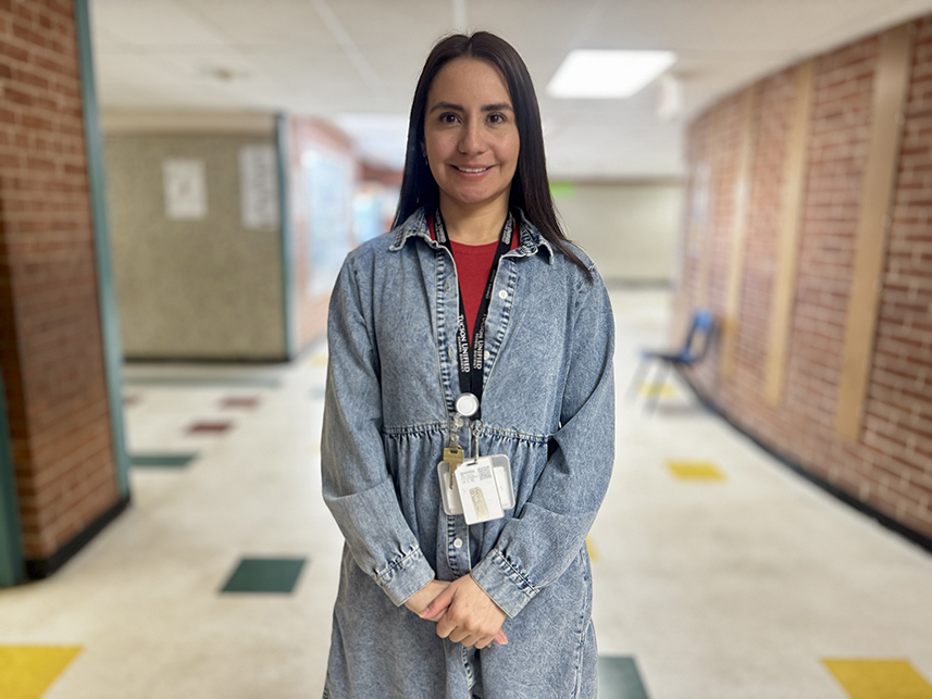 A woman with dark hair and a denim dress smiles in a school hallway